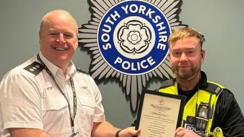 South Yorkshire Police A tall bald smiling man in a white shirt hands a framed certificate to a police officer with ginger hair and a beard, wearing a neon vest. They are both smiling at the camera.