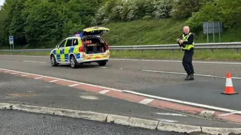 A police officer stands in the road on a section of the A1 between Morpeth and Berwick. A police car with an LED sign reading 'Road Closed' is parked in the road.
