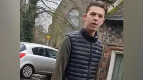 Norfolk Police Elvis Price is wearing a green jumper and black gillet. He is outside in wintery conditions and there is a silver car parked behind him.