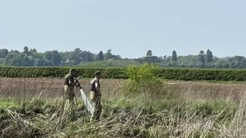 Two men in beige overalls standing in tall grass with a hedgerow in the distance