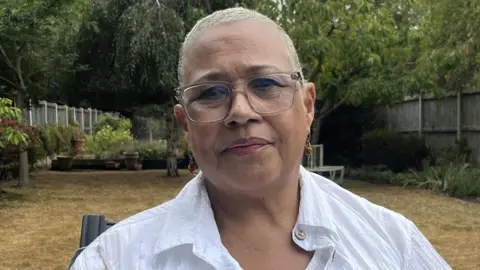 Mina Smallman, wearing a white blouse and glasses, sits in a garden. 
