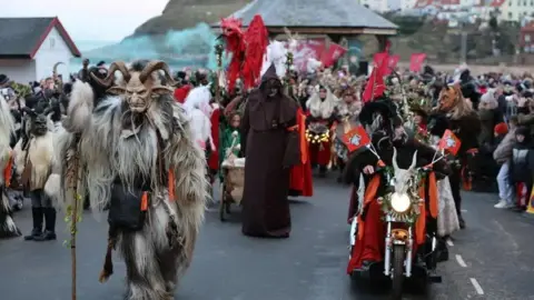 Baxter Photography People dressed up as a mythical horned beast called Krampus walk in a parade through Whitby, holding red and white flags. One person is riding a motorbike. 