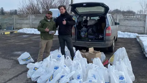 Ben Allen Two men stand behind a large collection of shopping bags on the floor in front of an open car boot. 