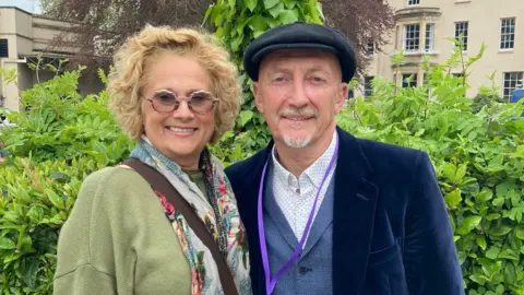Ian and Kim Holloway stand close together. Kim wears a green cardigan and pattern top. She has blonde wavy chin-length hair, and wears round glasses. She is smiling. Ian wears a blue velvet jacket, blue waistcoat, white open-necked shirt with spots. He has a grey/white goatee beard, and wears a flat cap on his head. He is smiling as he leans in towards his wife.
