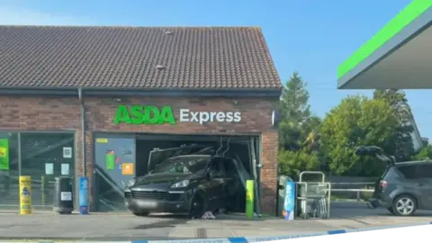 A Porsche Cayenne that is reversed into a shop window at an Asda Express petrol station. Police tape is across the forecourt. 
