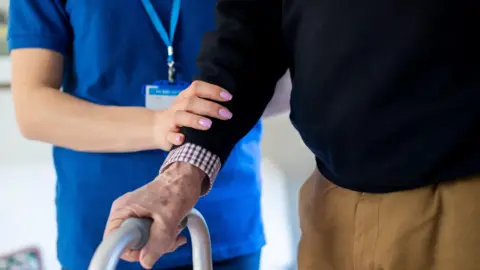 A care worker with her hand on an elderly man's arm, helping him to walk