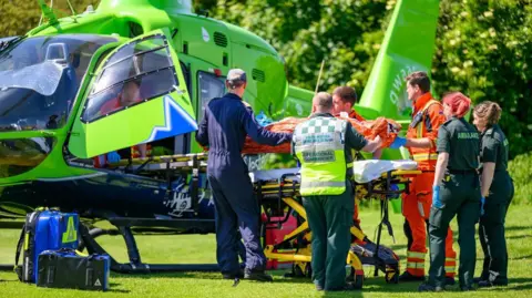 Paul Rubery Six crew members dressed in orange hi-vis suits, blue overalls, or dark green ambulance uniforms. They are loading a mannequin on a stretcher into a green air ambulance helicopter during a training exercise in the middle of a field.