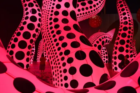 Giant luminous pink tentacle-like sculptures covered in black spots in a dark gallery, with visitors dwarfed by their scale