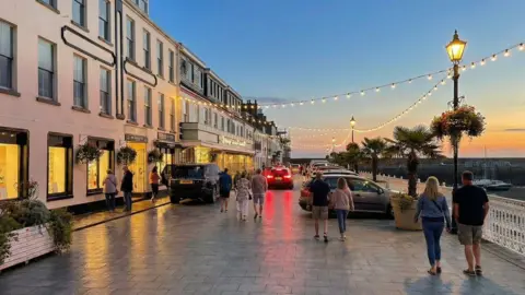 Gorey Pier Design Code This is an AI-generated image of Gorey Pier at sunset. The road is paved. People are walking down the road.