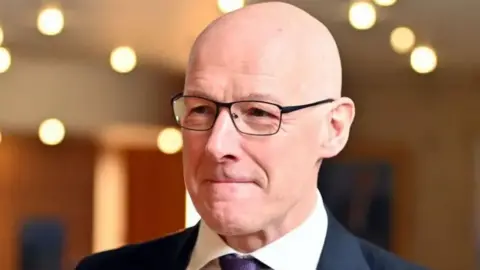 Getty Images Shaven-headed John Swinney in black glasses wearing a dark suit, white shirt and purple tie