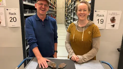 Leeds Museums and Galleries A smiling man and woman stood in front of a display trolley. On the trolley is a black sheet with the fossilised remains. 