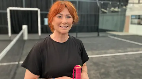 A woman dressed in a black T-shirt with red hair holds a padel racquet and stands on a court.
