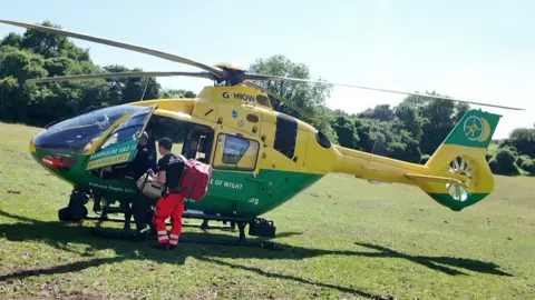 Yellow and green helicopter sitting on grass in a field, in front of trees and bushes, with one of its doors open and two men stood loading bags onboard