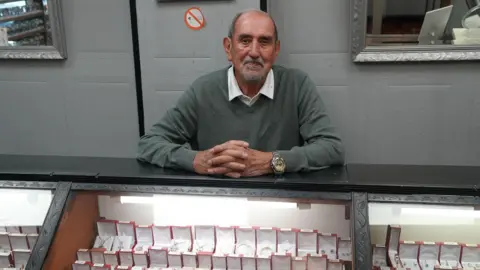 Jeweller Michael Newth is wearing a shirt and jumper and sitting behind his jewellery counter. 
