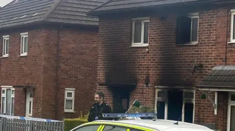 A police officer and police car are stood outside a property which has a number of burned out windows and door. There is fire damage to the brickwork above the windows and door.