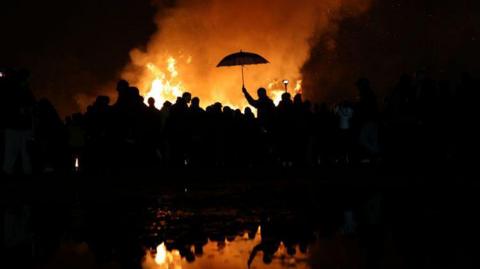 A crowd silhouetted in front of a bonfire, with a man holding up an umbrella