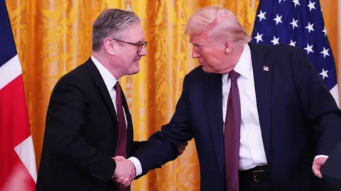 PA Media Sir Keir Starmer e Donald Trump apertam as mãos, sorrindo, na frente de suas respectivas bandeiras. Ambos os homens estão vestindo ternos escuros, camisas brancas e laços estampados em vermelho e azul.