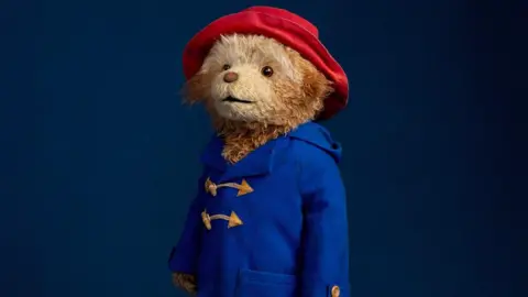 Jay Brooks The musical's Paddington bear stands wide-eyed in front of a dark blue background. He has a toy-like quality, wearing his famous blue duffle coat and large red hat.
