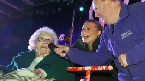 Norma is pictured switching on the Christmas lights at a shopping centre in Lincolnshire. She is speaking into a microphone held by a man in a purple jumper with a BBC Radio Lincolnshire logo