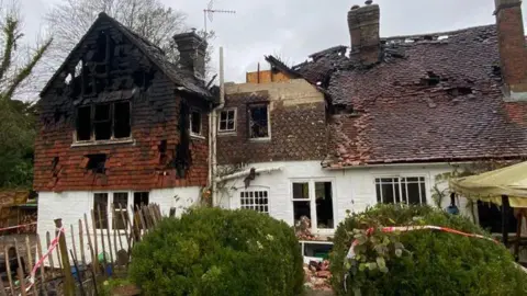 The image shows a house that has burned down, with holes in the roof and red and white safety tape around it and on the bushes at the front of the house. The sky in the background is grey and bushes can be seen on the left-hand-side.