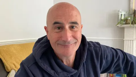 Nic Pioli is sat down inside his house, wearing a blue jumper.