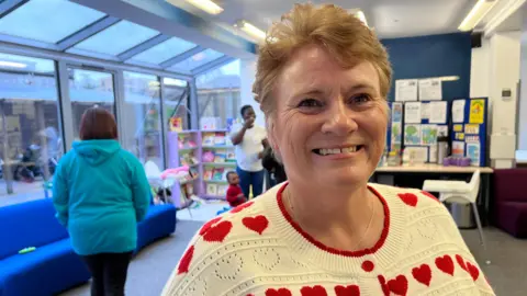 A lady wearing a cream coloured jumper, with a red heart design across the front and shoulders, smiles at the camera.