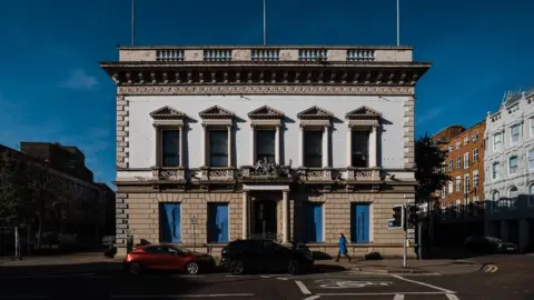 A white and brown building with columns at the sides of the windows. Cars are parked outside. A person wearing a blue coat is walking by. The sky is blue with minimal clouds. 