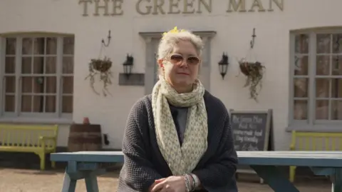 Shaun Whitmore/BBC Kerstin Rivett is sat on a blue picnic bench outside The Green Man pub. She is wearing sunglasses, a grey cardigan, silver bracelets and a yellow flower patterned scarf. She has white hair with daffodils tied into the back of her hairdo. 