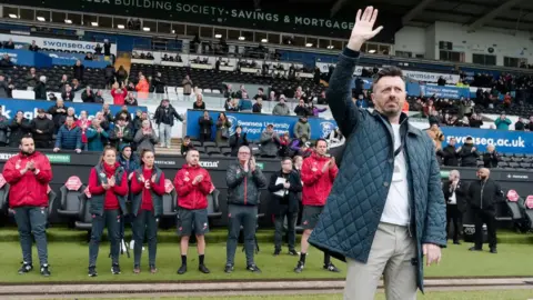Swansea City AFC Jason Bowen in a padded jacket and beige trousers waving at the side of a football pitch, as coaches and fans in the stand behind him applaud