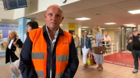 Loïc Andro, who is wearing an orange and black high-visibility vest,  stands in an indoor public space. In the background, several other people are walking or standing, some carrying bags. There is a digital display screen showing information and a sign that reads Welcome to Jersey.