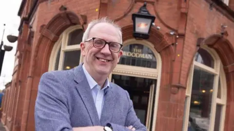 A photograph of a man wearing glasses stood outside a traditional pub with "The Woodman" written on glass above the door behind him. He has his arms crossed and is wearing a suit jacket and shirt. 