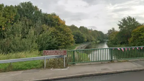 A pavement on a bridge with a brown sign reading "Bridge Street" with "Clayhanger Road" below, and green railings to the right. There is a canal under the bridge and a grass area and trees to the left, and more trees to the right.