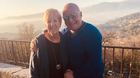 Aldo Zilli Aldo Zilli, who is a bald man wearing a blue V-neck jumper and sunglasses, has his arm around his mother-in-law Maureen, who has short blonde hair and is wearing a zipped hoodie over a black top and a long necklace. They are both standing in front of a hilly backdrop and are smiling at the camera.