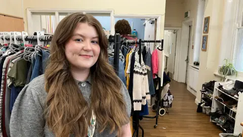 A woman with long brown hair in a grey button up jumper stands in front of two clothing rails with lots of different tops and jumpers on them. On the right hand side, two shoe racks can be seen. 