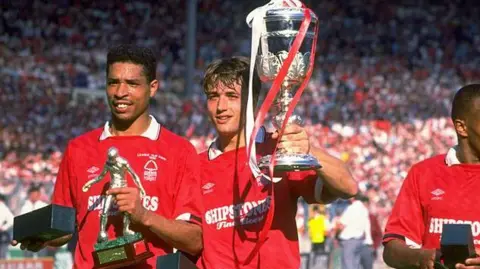 Getty Des Walker (left) and Nigel Jemson in a red strip on the pitch at Wembley. Des is holding a silver figure of a footballer in his left hand and Nigel is holding up the Littlewoods cup in his left hand, which has red and white ribbons trailing off it. Behind them are banked rows of supporters in the stands. 