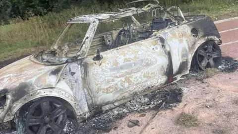 Richard Whitehouse A burnt-out Mini car sitting on a cinder running track. The tyres have been completely destroyed. The wheel rims are fully blackened. The car has been stripped of all its paint and has lost its roof.