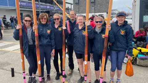 Truro River Rowing Club Six gig rowers standing on land holding their oars in the air.