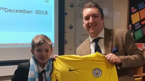 Painsley Catholic College A boy in a school uniform and striped scarf stands next to a man, Adam Reynolds, who is holding a yellow football shirt. They are both standing in a classroom setting