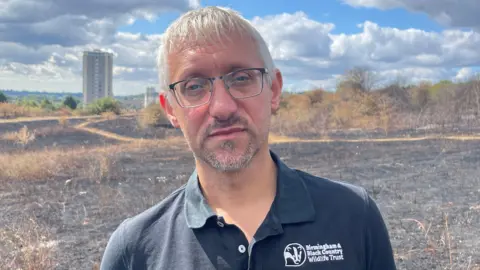 A man with white-grey hair and black glasses. He is wearing a black t-shirt that has a badger logo on the breast and says "Birmingham & Black Country Wildlife Trust". Behind him is a field in a nature reserve completely blackened and charred by fire. There are very few pathway shapes not affected by fire, and they are light brown dead grass. In the distance are high rise buildings