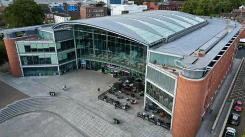 A drone image of The Forum building. There is a glass atrium and a curved main roof, with flatter roofs around the sides of the U-shaped building. The BBC office is in the left-hand wing. There is a paved area, stone steps as well as tables and chairs outside the building.