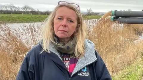 Bryony Sadler has long blonde hair and is pictured outside in Somerset. She is wearing a navy blue coat with a grey fluffy lining and a green scarf. She has glasses on her head. Behind her are pumps, pumping flood water out.