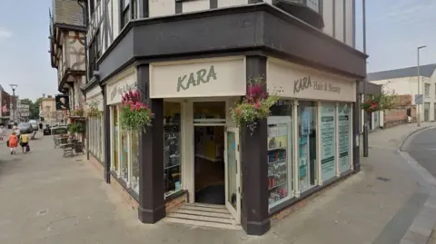 Google A screenshot from Google street view showing the entrance to Kara Hairdressers. It is positioned on the corner of a junction with an open door and concrete pillars either side. The building is Tudor style with wooden beams across the exterior. The frontage is painted light brown with dark brown accents and hanging flowers baskets outside. In the window there is a display of hair products.