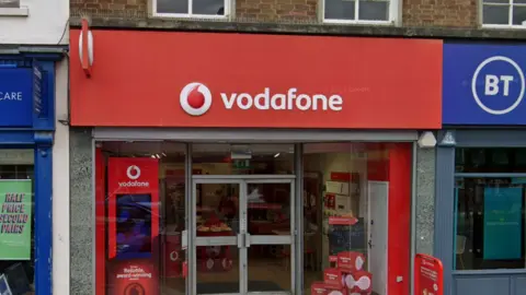 The outside of the Vodafone shop on the High Street in Newmarket. It has a red sign saying "vodafone" over the door and glass windows and doors. 