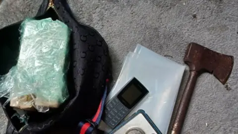 A bag with a package in green plastic, a mobile phone, some plastic bags and a small axe