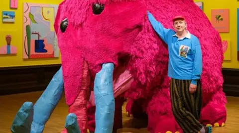 Tom Bird A man in striped black and beige trousers stands his arm resting on a life-size sculpture of a woolly mammoth, with pink fur, blue horns and yellow toenails. He is in an art gallery with yellow walls, with paintings hung.
