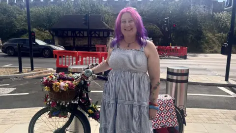 A woman with pink and purple ombre hair and pale skin stands in front of a cordoned off cycle lane with her bicycle. She is wearing a navy and white stripe sundress, and blue and red bangles on her wrist. Her bicycle is black with a basket on the front and fake flowers and fairy lights twined around it.