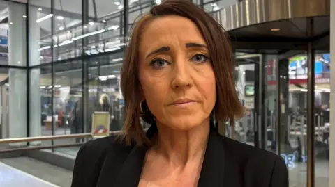A woman with short dark hair in a black jacket standing in front of glass doors looks directly at the camera 