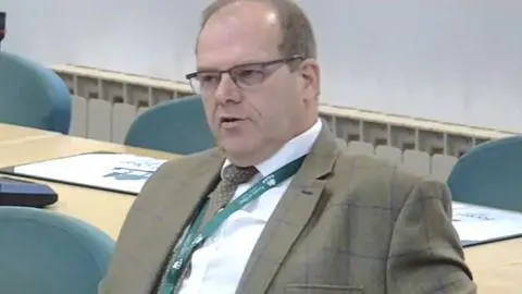 Man in a green suit and white shirt. He sits back in a chair in an indoor setting with a table behind him. He also wears a lanyard.