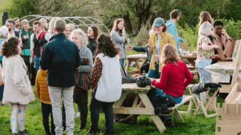 Ibolya Feher Knowle West Spring Celebration - shows groups of people standing and sitting on benches, eating and drinking together in a field