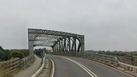 Iron bridge over a two lane road with double white lines in the middle of the road. There is a pavement on either side of the bridge 